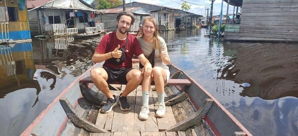 Two people sitting in a wooden boat on a calm river with houses on the opposite bank. 1