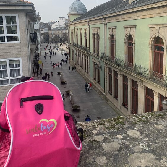 A pink backpack with 'sintelugo free tour' on it, overlooking a historic square with people walking, buildings with arches, and a dome structure in the background. 2