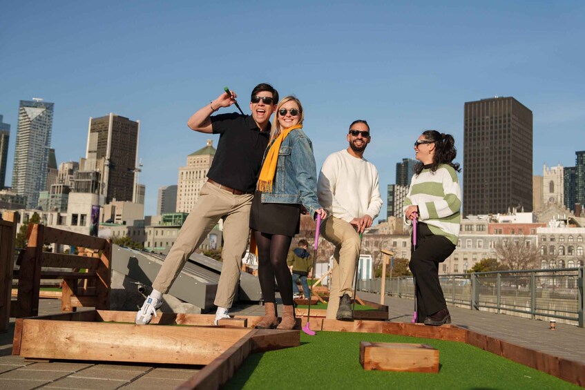 Eine Gruppe von Menschen spielt Minigolf auf einem Dach, im Hintergrund ist eine Stadtansicht zu sehen. 1