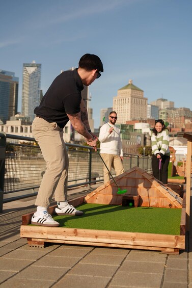 Eine Person spielt Minigolf auf einem Außenplatz, im Hintergrund ist eine Stadtlandschaft zu sehen. 2