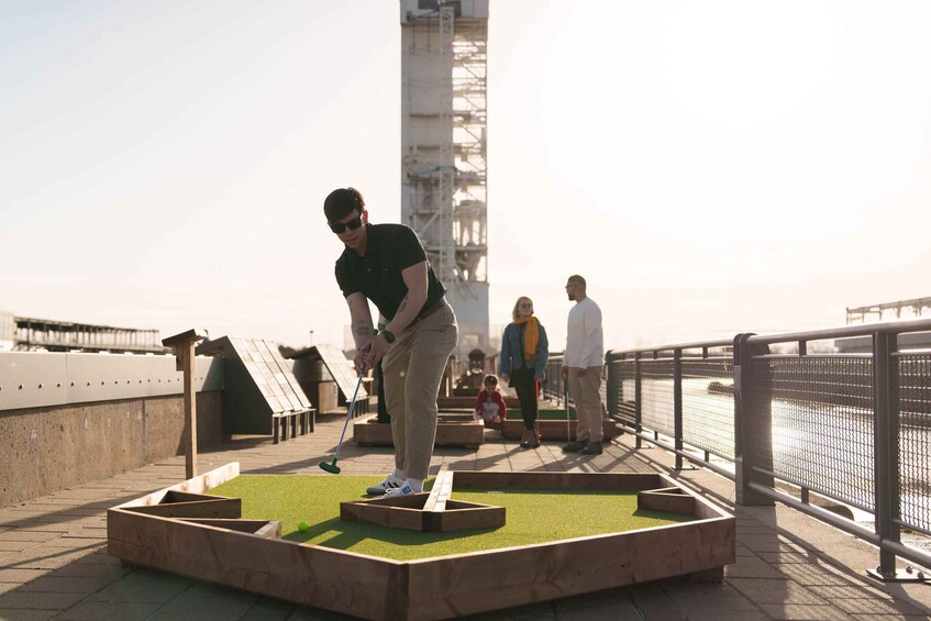 Eine Person spielt Minigolf auf einem Dach, im Hintergrund ist ein Gewässer zu sehen. 3
