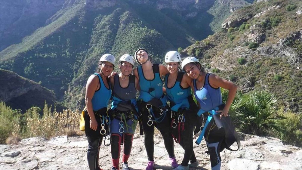 A group of people wearing helmets and harnesses, standing on a rocky terrain with a mountainous landscape in the background. 5