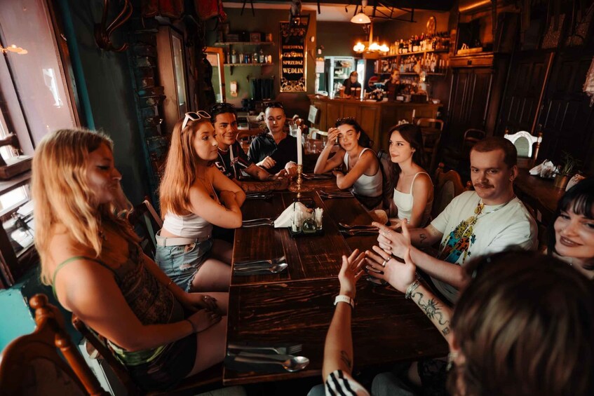 A group of people seated around a wooden table in a dimly lit bar with shelves of bottles in the background. 2