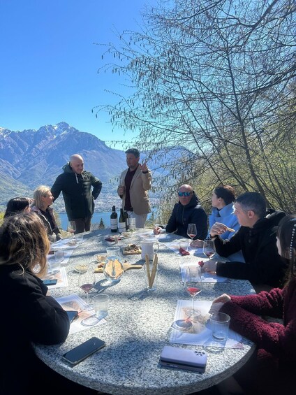 A group of people seated around a table outdoors with a mountainous landscape in the background. 5