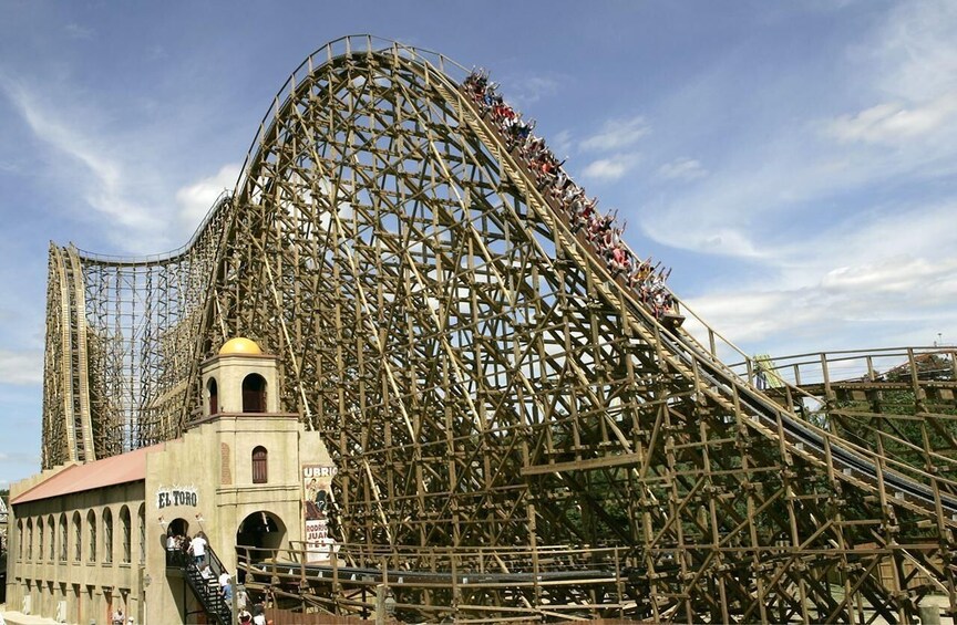A wooden roller coaster with a steep drop and a station with a bell tower. 2