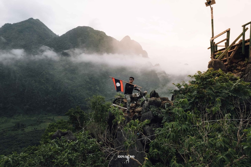 A person standing on a rocky outcrop with a flag, surrounded by lush greenery and misty mountains in the background. 5