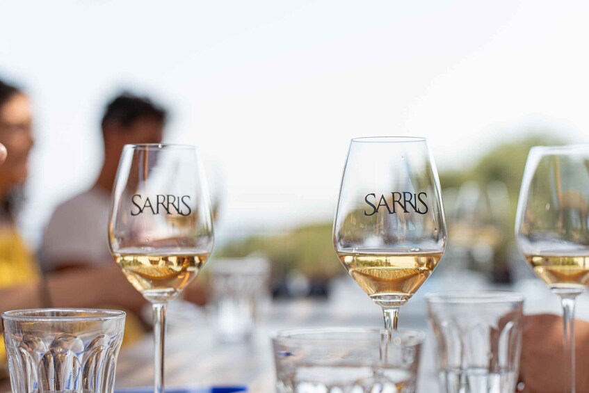 A row of wine glasses with the name 'SARRIS' on them, filled with white wine, placed on a table. 1