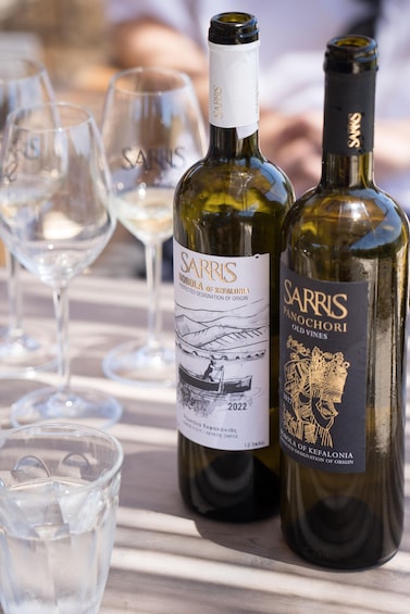 Two bottles of Sarris wine, one labeled 'Robola of Kefalonia' and the other 'Panochori Old Vines', on a table with empty wine glasses. 2