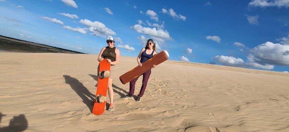 Two people in a desert with sand dunes, one holding a snowboard. 1
