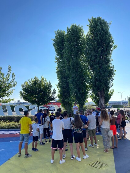 A group of people gathered outdoors, possibly at a public event or gathering, with trees and a clear sky in the background. 3