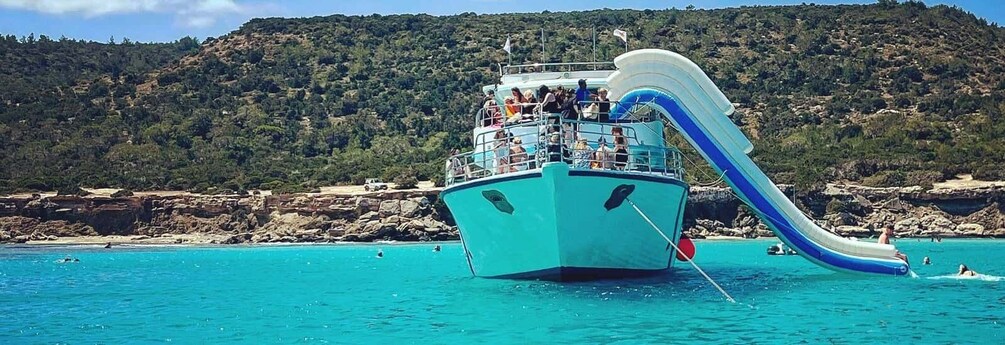 A boat with a water slide in turquoise waters, surrounded by rocky cliffs and a forested hill. 4
