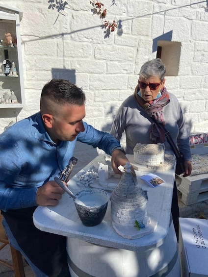 Two people working on a stone structure outdoors, with a white brick wall in the background. 2