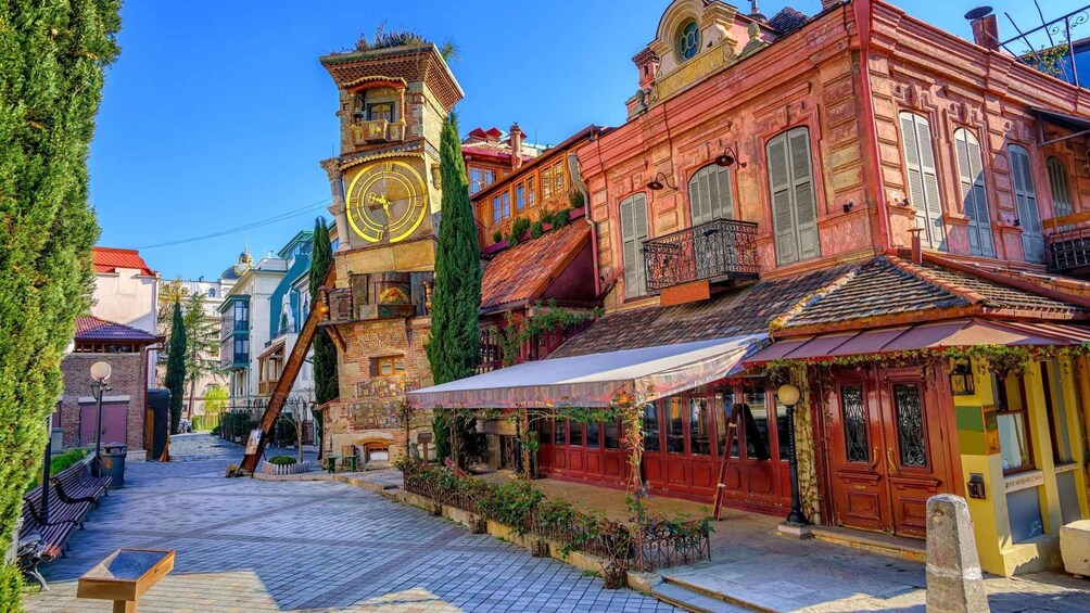 A cobblestone street lined with colorful buildings, a clock tower, and a café with outdoor seating. 2