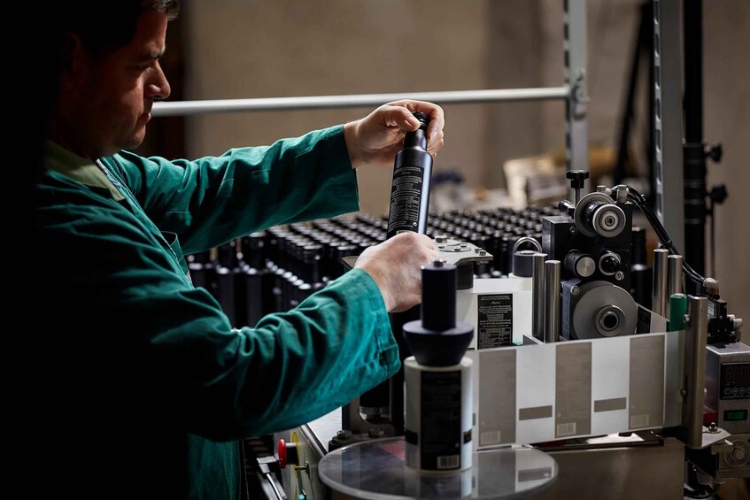 A person in a green lab coat handling a black bottle in a laboratory setting with various scientific equipment. 2