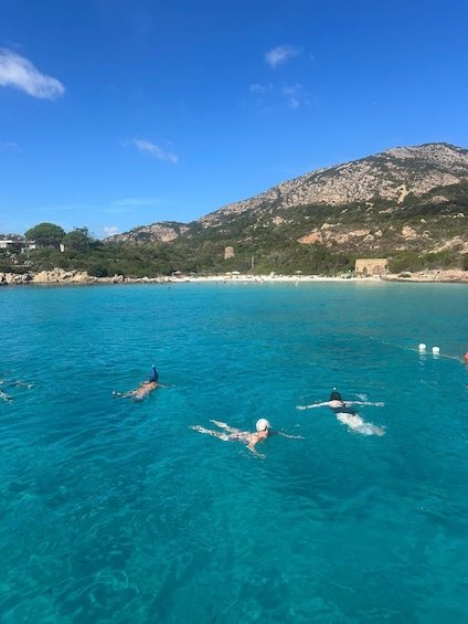 A clear blue sea with people swimming and snorkeling. In the background, there are rocky hills and a few buildings. 3