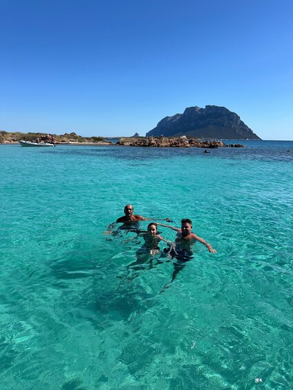 Three people snorkeling in clear turquoise water with a rocky coastline and a prominent mountain in the background. 4