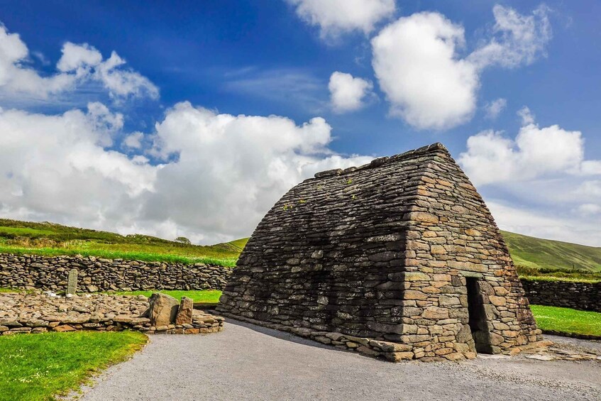 A stone structure with a triangular roof, surrounded by a stone wall and grassy fields under a partly cloudy sky. 2