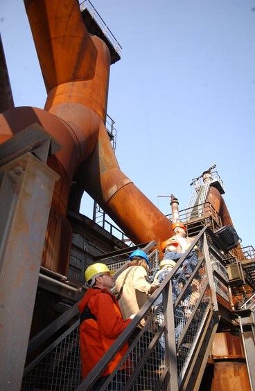 Workers in safety gear climbing a metal staircase next to large industrial pipes. 3