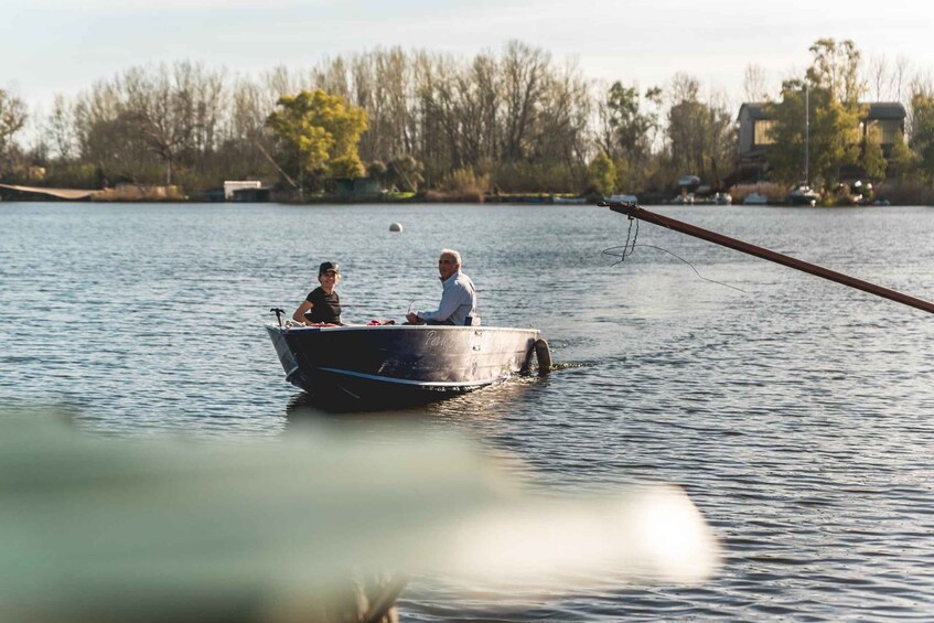 A boat on calm water with two people, surrounded by trees and buildings in the background. 1