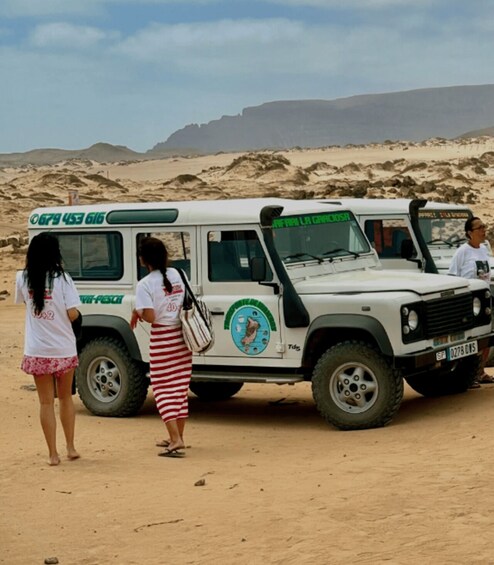 A white safari vehicle with a logo and text on its side, parked on a sandy terrain with a desert landscape in the background. 2