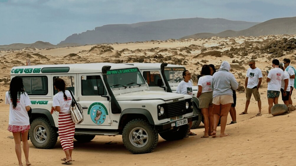A group of people standing around a white vehicle with a logo and text on its side, parked on a sandy beach with mountains in the background. 1