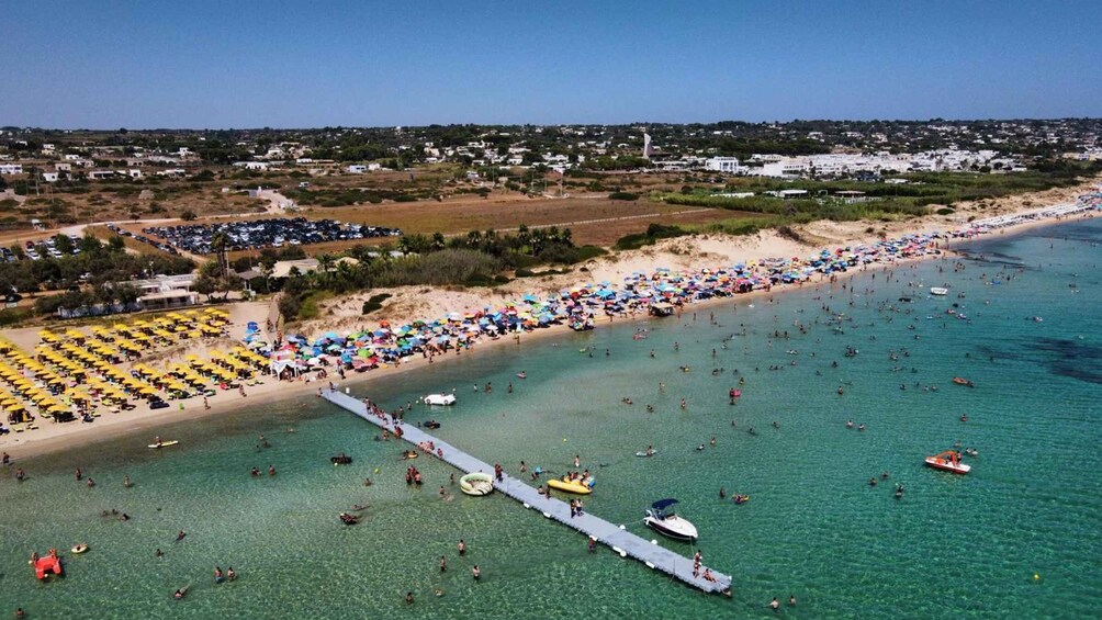 A crowded beach with people swimming and sunbathing, a pier extending into the water, and a distant town. 5