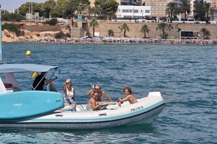 A small boat with three people on a calm sea, a beach with people in the background, and buildings along the shore. 4