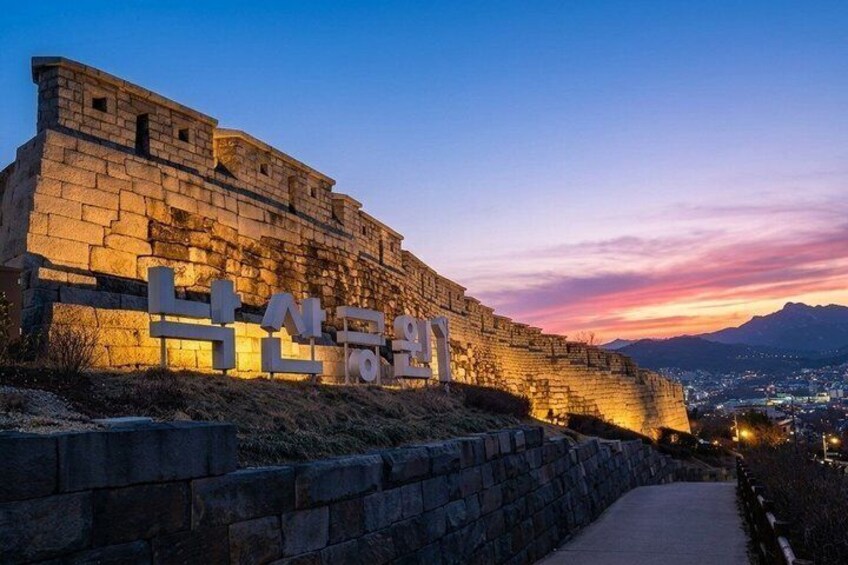 A stone wall with illuminated Korean characters, a pathway, and a sunset in the background. 1