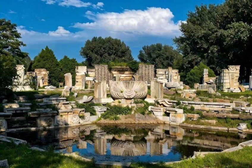 Ancient ruins with columns and a central fountain, surrounded by trees and a body of water. 4