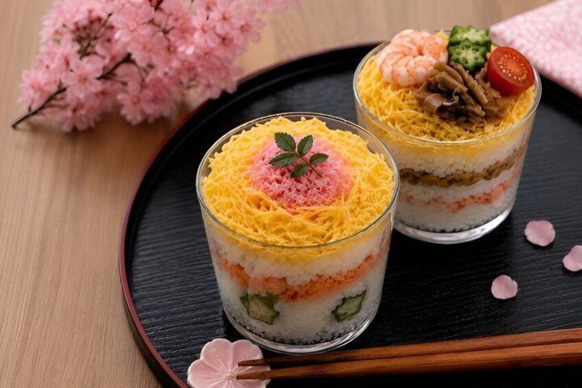 Two layered desserts in clear glasses, garnished with pink foam, shrimp, and vegetables, on a black tray with cherry blossoms in the background. 1