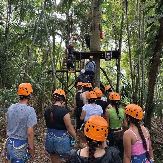 A group of people wearing helmets and harnesses, preparing for a zip-lining activity in a forested area. 2