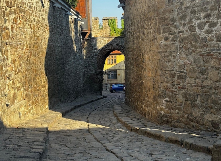 A narrow cobblestone street flanked by stone walls, leading to a yellow building with a red-tiled roof. 2