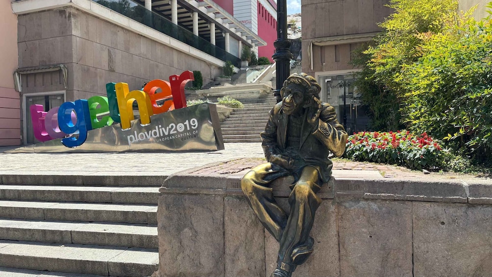 A statue of a seated figure on a stone bench with a backdrop of a building and a sign reading 'Together'. 2