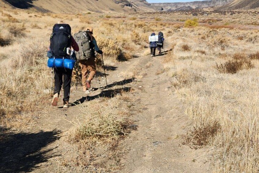 A group of hikers on a dirt trail in a dry, grassy landscape with hills in the background. 5