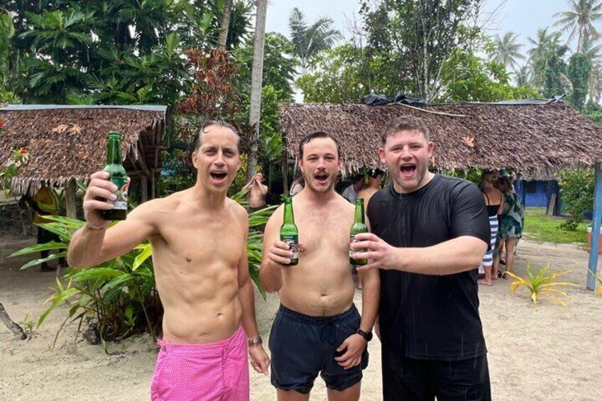 Three people holding bottles, a thatched-roof hut, and tropical vegetation. 4