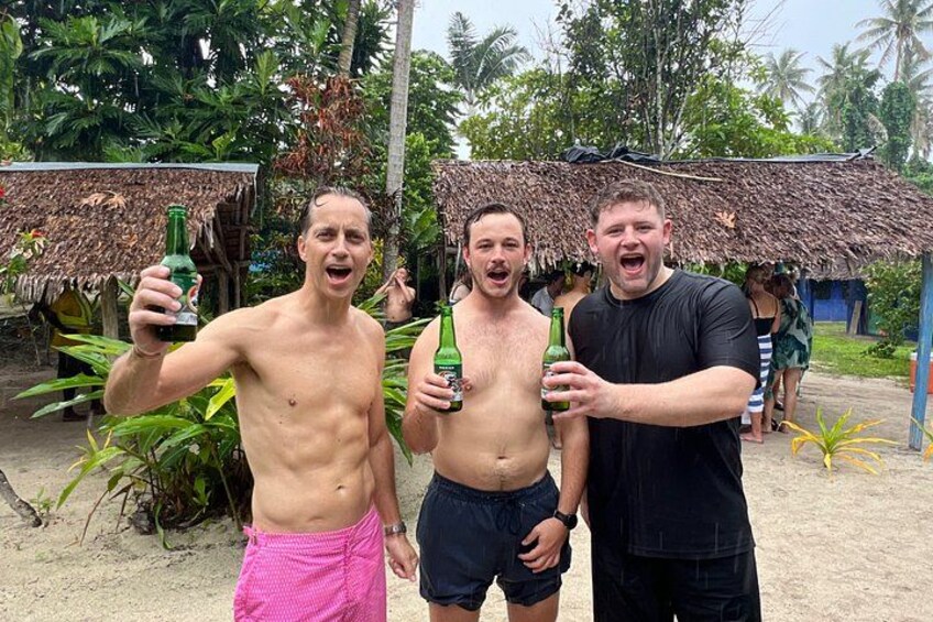 Three people holding bottles, a thatched-roof hut, and tropical vegetation. 2
