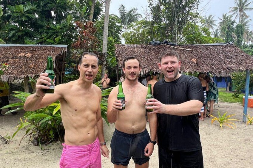 Three men holding bottles, standing in a tropical setting with thatched-roof huts and palm trees. 4