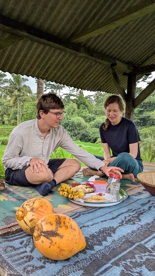 Two people sitting on a mat under a shelter, with food and drinks in front of them. 5
