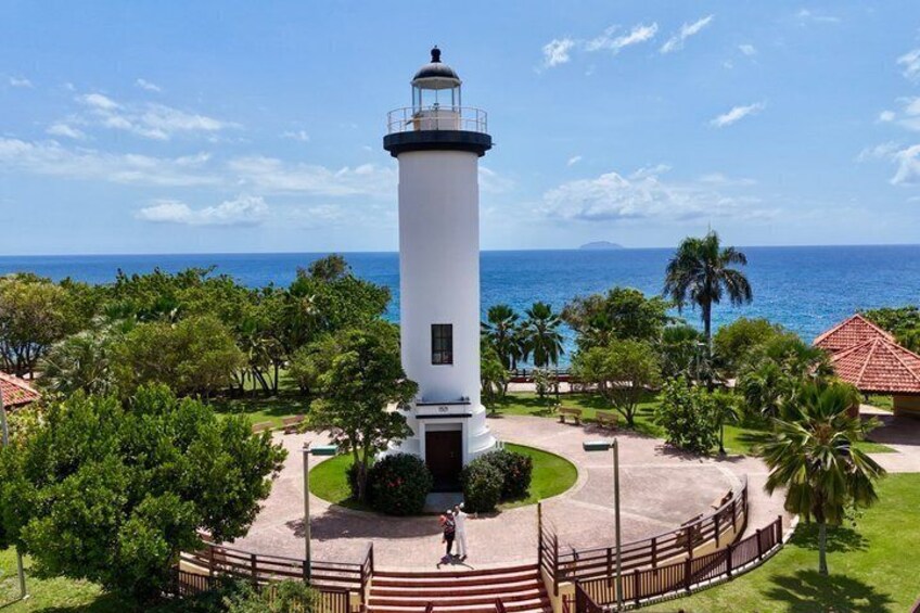 A white lighthouse with a black top, surrounded by a circular walkway, palm trees, and a view of the ocean. 1