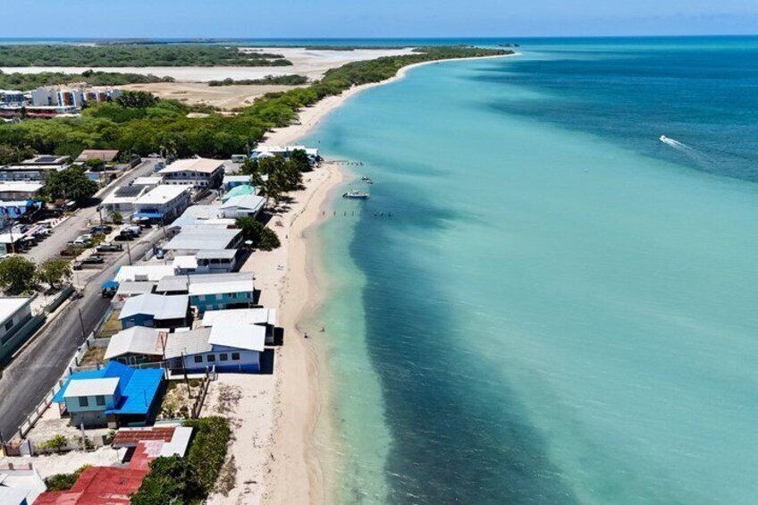 A coastal area with a sandy beach, clear turquoise waters, and a row of buildings along the shore. 2