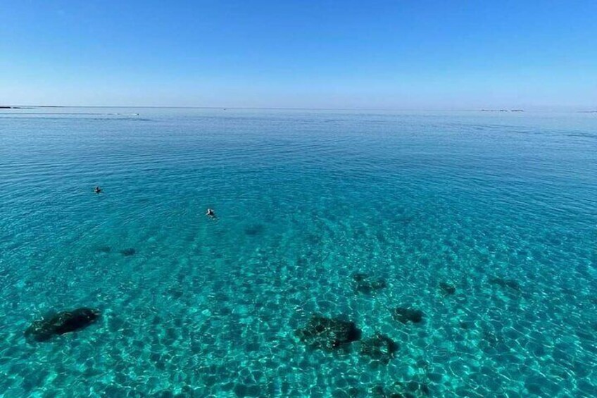 A clear blue sea with visible underwater rocks and a few people swimming. 2