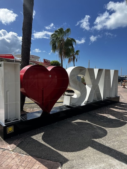 A large heart-shaped structure with the letters 'SXIV' next to it, set against a clear blue sky with palm trees in the background. 4