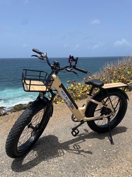 A beige electric bike with a basket and a rear rack parked on a gravel path by the ocean. 3