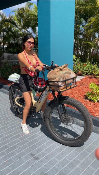 A person sitting on a bicycle with a basket, surrounded by greenery and a blue structure. 2