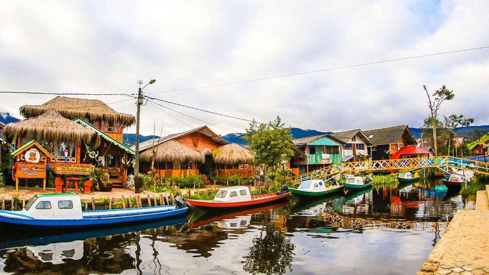 A riverside village with thatched-roof houses, a bridge, and boats docked along the water. 2