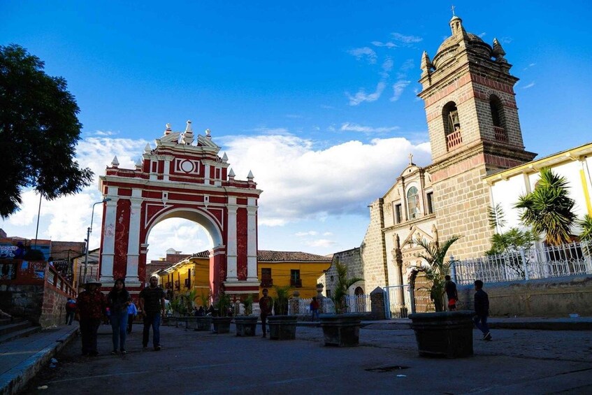 A historic archway with a clock, a church with a bell tower, and a plaza with people and palm trees. 3