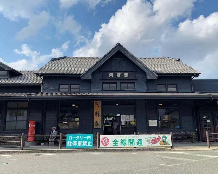 A building with a dark facade and a sign that reads 'Arashiyama.' There are two signs in front: one with a red arrow and the text 'Rooftop parking' and another with a green background and the text 'JR Arashiyama Station.' 2