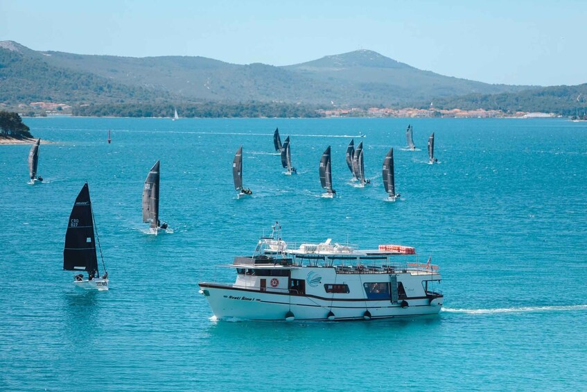 A sailing regatta with multiple sailboats on a calm sea, a larger boat with a cabin, and a mountainous coastline in the background. 1
