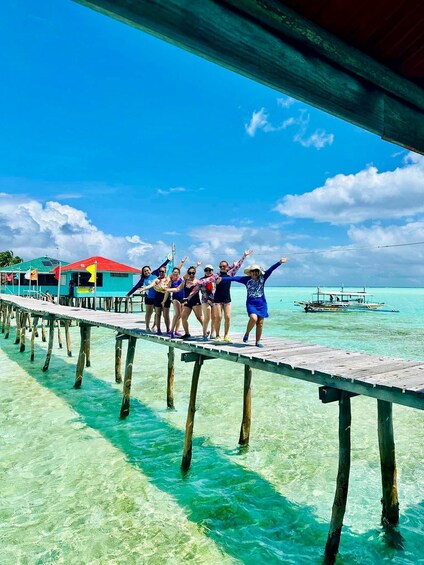 A group of people on a wooden pier extending into turquoise water, with a boat and buildings in the background. 1