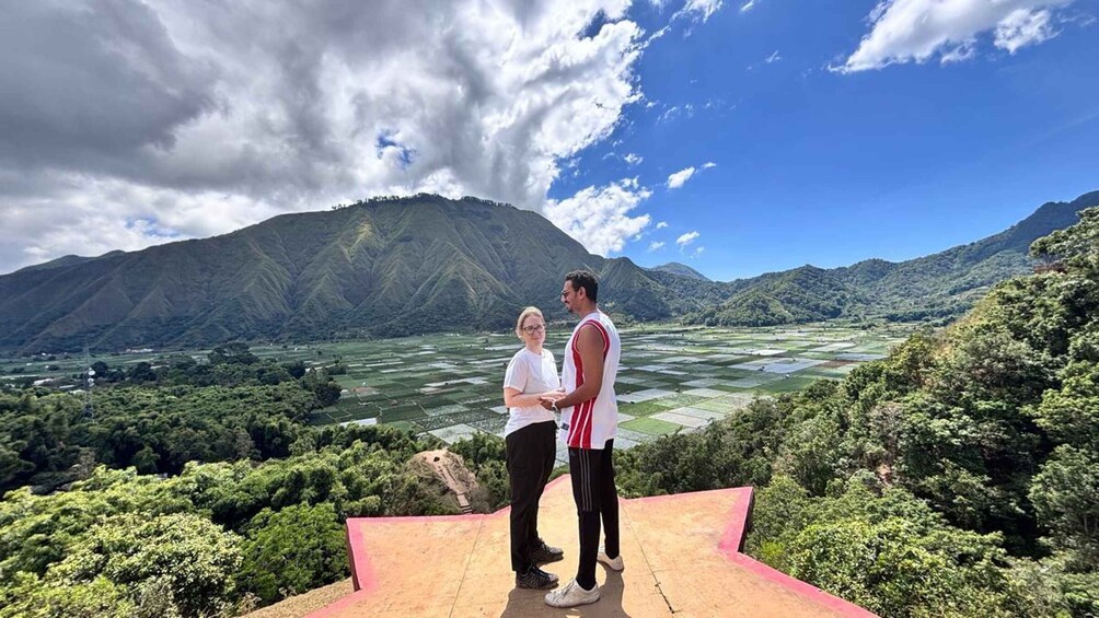 Two people standing on a platform overlooking a mountainous landscape with a river and fields. 1